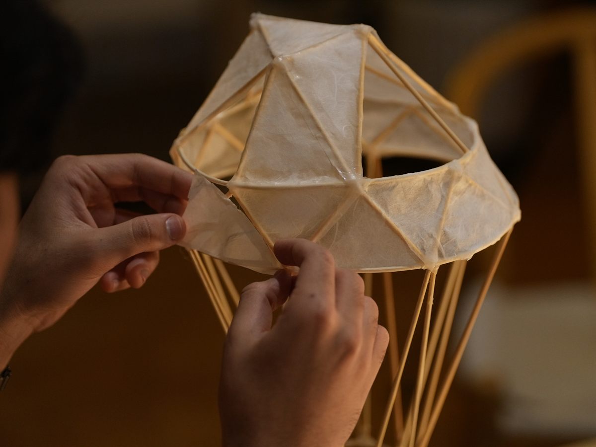 Hands attaching a paper panel to a geodesic bamboo lamp frame in a darkened room.