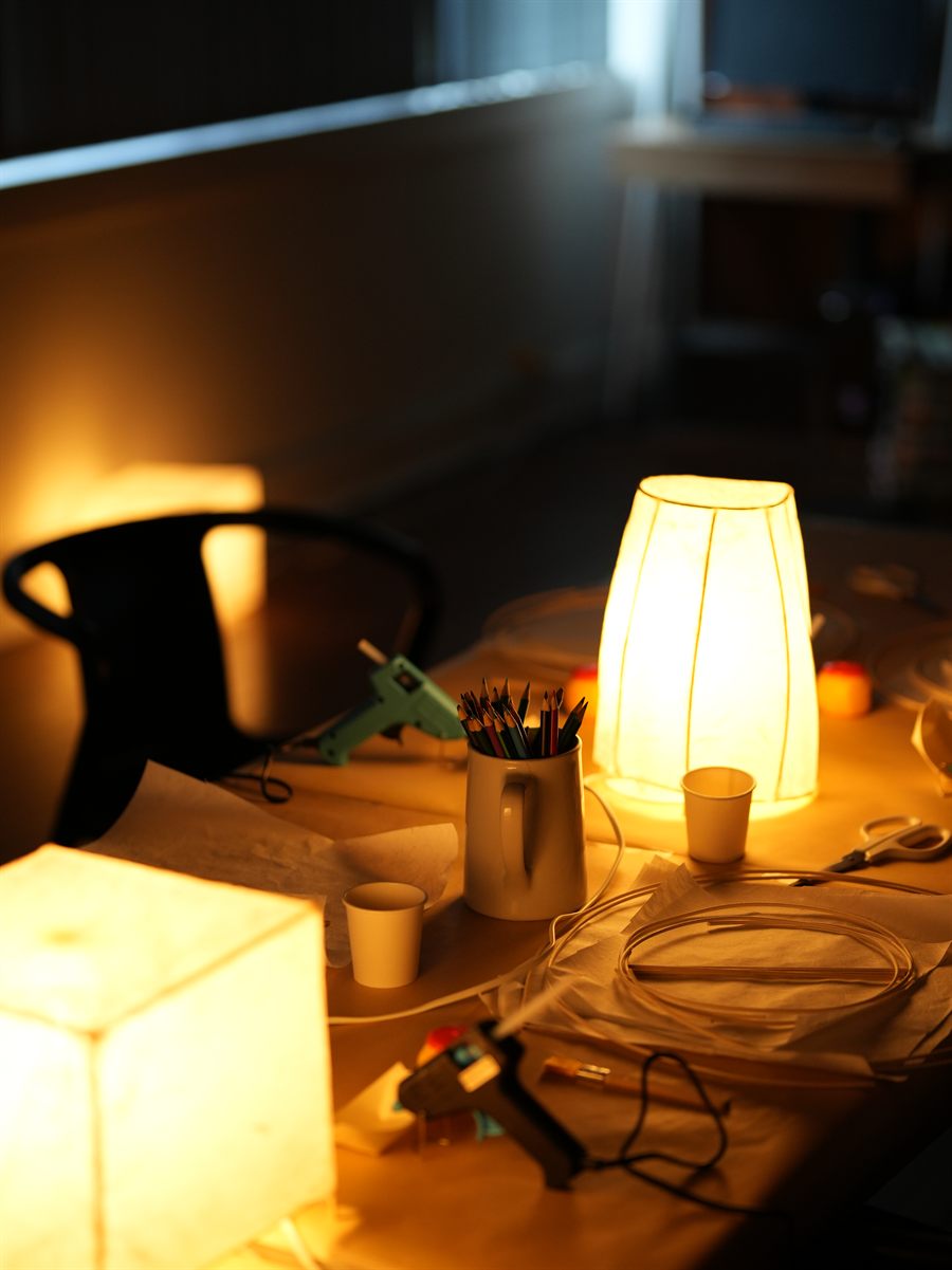 Two finished paper lamps glowing on the workshop table among pencils, tools and offcuts.