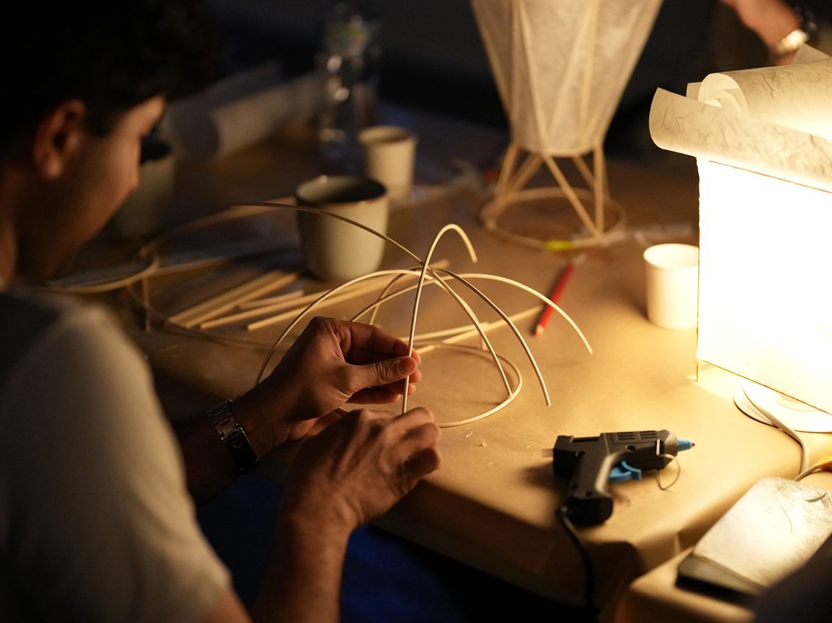 A workshop participant bending bamboo cane into a curve beside a glowing paper lamp.
